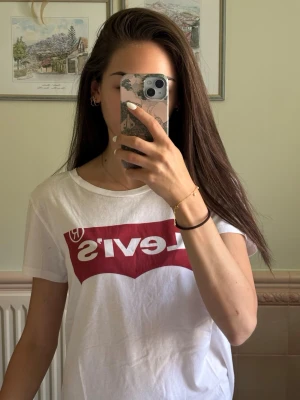 Vit Levi's t-shirt med röd logga - Vit t-shirt från Levi's med klassisk röd logotyp framtill. T-shirten har rund halsringning och korta ärmar. Tillverkad i mjuk bomull, perfekt för en avslappnad och stilren look.