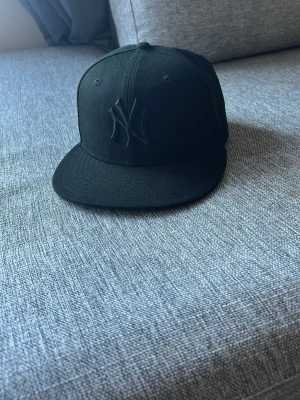Svart New Era Yankees keps - Säljer en svart New Era 59FIFTY keps med New York Yankees-logga i svart brodyr framtill. Klassisk flat peak och strukturerad passform. Perfekt för dig som gillar stilrena accessoarer och sportig look.