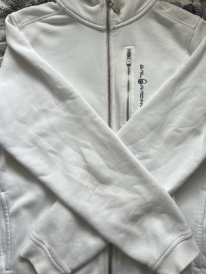 Vit hoodie från Sail Racing - Vit hoodie från Sail Racing med dragkedja framtill och en extra dragkedjeficka på bröstet. Insidan är fodrad med mjuk fleece för extra komfort. Snygg logga på bröstet och huva med dragsko. Perfekt för en sportig och avslappnad stil.