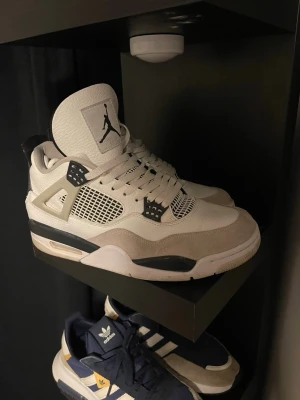 Vita Air Jordan 4 sneakers - Snygga Air Jordan 4 sneakers i vitt med grå och svarta detaljer. Skorna har klassisk meshpanel på sidorna, synlig Air-sula och Jumpman-logga på plösen. Ovandelen är i läder och mocka för en stilren look.