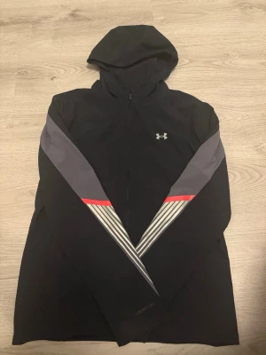 Svart vind jacka från Under Armour - Snygg svart hoodie från Under Armour med dragkedja framtill och huva. Ärmarna har gråa och vita detaljer samt en röd linje som ger en sportig look. Tillverkad i ett tekniskt material som andas, perfekt för träning eller chill.