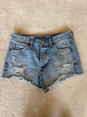 Ljusa blå jeansshorts med slitningar - Säljer ett par ljusblå jeansshorts med råa kanter och slitna detaljer framtill. Klassisk femficksmodell med bälteshällor och knappgylf. Perfekta för sommaren och ger en avslappnad vibe.