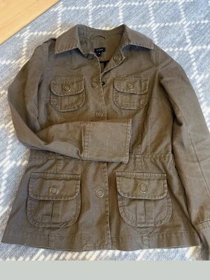 Brun utilityjacka från Lindex - Snygg brun utilityjacka från Lindex med fyra stora fickor framtill och klassisk krage. Jackan har knappar och är tillverkad i ett slitstarkt bomullsmaterial. Perfekt för lager-på-lager och en cool vardagslook.
