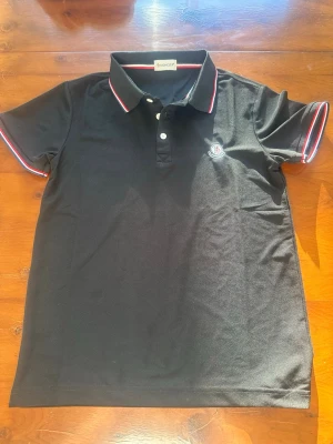 Svart moncler piké - Snygg svart pikétröja med klassisk krage och knappar framtill. Tröjan har röda och vita detaljer på krage och ärmslut samt en liten logga på bröstet. Perfekt till en avslappnad men stilren look.