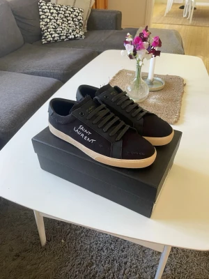 Svarta Saint Laurent sneakers - Snygga svarta sneakers från Saint Laurent med vit logga på sidan. Skorna har svart ovandel i canvas och läderdetaljer, samt en beige sula som ger en clean kontrast. Klassisk låg modell med svarta skosnören och diskreta detaljer.
