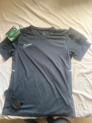 Svart Nike sporttröja - Snygg svart sporttröja från Nike med klassisk vit swoosh-logga på bröstet. Tröjan har korta ärmar och är tillverkad i ett lätt och snabbtorkande material som passar perfekt till träning eller match. Enkel och stilren design med små vita detaljer på sidorna.Pris kan diskuteras.
