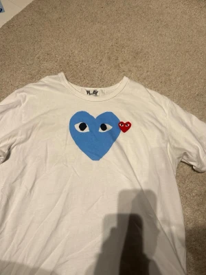 Vit Comme des Garçons PLAY t-shirt - Säljer en vit t-shirt från Comme des Garçons PLAY med ett stort blått hjärta med ögon och ett litet rött hjärta på bröstet. Klassisk rund hals och korta ärmar. Tillverkad i mjuk bomull, perfekt för en avslappnad och trendig stil.