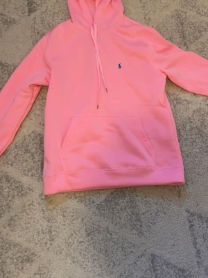 Rosa hoodie med huva från Polo Ralph Lauren - Säljer en snygg rosa hoodie från Polo Ralph Lauren med klassisk huva och dragsko. Tröjan har en stor magficka och den ikoniska lilla blå loggan broderad på bröstet. Perfekt för dig som gillar streetwear och vill ha något bekvämt och stilrent.