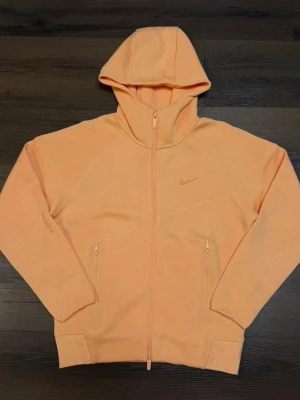 Hoodie från Nike NOCTA - Säljer en persikofärgad/orange hoodie från Nike NOCTA med dragkedja framtill och två fickor med dragkedja. Hoodien har en stilren design med liten Nike-logga på bröstet och en skön huva. Materialet är mjukt och stretchigt, perfekt för chill dagar.