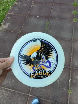 Innova Star Eagle discgolf disc - Säljer en vit discgolf disc från Innova, modell Star Eagle. Discen har en stor illustration av en örn med utbredda vingar och en orm, samt texten 'Star Eagle' och siffrorna 7/4/-1/3. Perfekt för discgolfentusiaster.