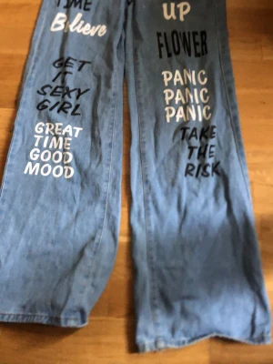 Blå jeans med tryck och vida ben - Unika blå jeans med breda ben och coola tryck i svart och vitt på framsidan, bland annat texter som 'Believe', 'Panic Panic Panic' och 'Take the risk'. Jeansen har en avslappnad passform och är tillverkade i klassisk denim. Perfekta för dig som vill sticka ut med din stil.