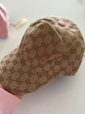 Beige keps med GG-mönster  - Lapp kvar, helt ny och oanvänd. Snygg beige keps från GG med det klassiska GG-monogrammet över hela kepsen. Kepsen har en böjd skärm och en rosa knapp på toppen som ger en unik touch. Tillverkad i slitstarkt tyg, perfekt för en trendig streetwear-look.