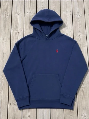 Mörkblå hoodie från Polo Ralph Lauren - Snygg mörkblå hoodie från Polo Ralph Lauren med klassisk röd logga broderad på bröstet. Hoodien har huva, känguruficka och ribbade muddar vid ärmslut och midja. Perfekt för en chill och avslappnad stil.