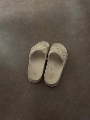Beige flätade slip-in sandaler - Säljer ett par beige slip-in sandaler med bred, flätad rem över foten. Sandalerna har en chunky sula och är tillverkade i ett mjukt material som ger en trendig och bekväm look. Perfekta för sommaren och lätta att matcha med olika outfits.