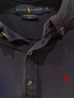 Blå skjorta från Ralph Lauren - Klassisk marinblå skjorta från Ralph Lauren med röd broderad logga på bröstet. Skjortan har långa ärmar, knappar framtill och button-down krage. Tillverkad i ett lätt och luftigt bomullsmaterial som passar perfekt till många olika stilar.