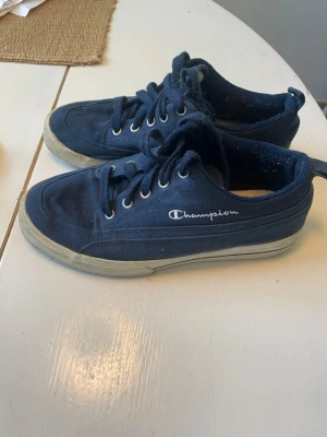 Mörkblå Champion sneakers i canvas  - Mörkblå sneakers från Champion med lågt skaft och klassisk snörning. Skorna har en vit sula och är tillverkade i canvasmaterial. Champion-loggan syns broderad på sidan. Perfekta för en avslappnad och sportig stil. (Katt finns) 