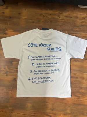 Maison de Rêve tshirt  - Vit t-shirt med blå text och tryck både fram och bak. På ryggen står det 'Côte d’Azur Rules' med roliga regler i blått. Klassisk passform och rund hals. Perfekt för dig som gillar stilrena plagg med en twist. Från maison de Rêve, helt ny men för liten för mig. Har kvitto och pris går att diskutera 