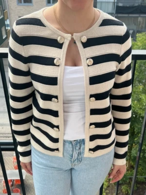 Randig kofta med pärlknappar - Snygg kofta med breda svarta och beige ränder, rund halsringning och dekorativa pärlknappar framtill. Koftan är stickad och har långa ärmar, perfekt att slänga över en t-shirt. Passar dig som gillar klassisk och trendig stil.