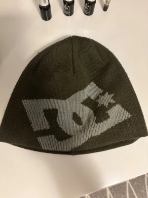Olivgrön DC mössa med logga - Säljer en olivgrön mössa från DC med stor grå logga framtill. Mössan är stickad i mjukt material och har en klassisk, rundad form som sitter skönt på huvudet. Perfekt för dig som gillar streetwear och vill ha en snygg accessoar till hösten och vintern.