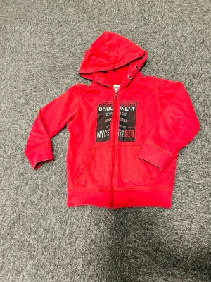 Röd hoodie med tryck från D.M.D - Cool röd hoodie från D.M.D med svart och vitt tryck på bröstet där det står Brooklyn och NYC. Dragkedja framtill, två fickor och skön huva. Materialet känns mjukt och bekvämt.