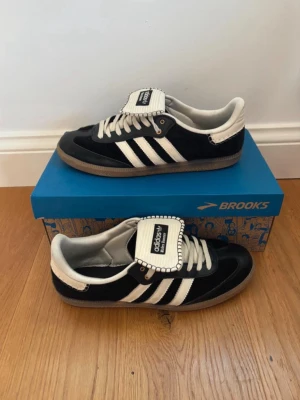Adidas svartvita sneakers med gummisula - Snygga Adidas sneakers Wales bonner i svart mocka med vita detaljer och klassiska tre ränder på sidan. Skorna har en bred vit plös med Adidas-logga och vita skosnören. Yttersulan är i gummi och ger en retrovibe.