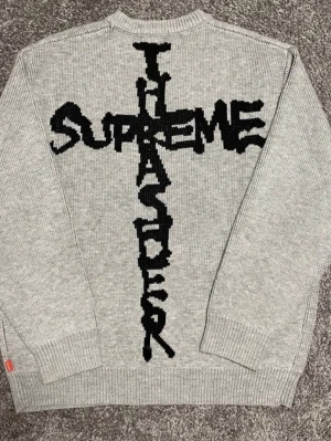 Grå stickad Supreme x Thrasher tröja - Stickad grå tröja från Supreme i samarbete med Thrasher. Tröjan har ett stort svart grafiskt tryck på ryggen med texten 'SUPREME' och 'THRASHER' i kors. Rund halsringning och ribbade muddar vid ärmslut och nederkant. Perfekt för streetwear-stilen. Använd en gång och är i storlek S. Priset är förhandlingsbart.