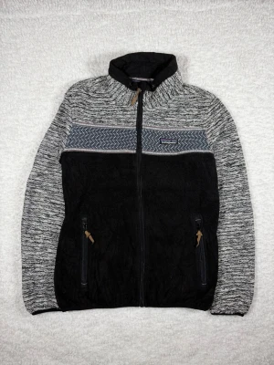 Svart och grå teddyjacka Patagonia - Mysig teddyjacka från Patagonia i svart och grått med mönstrad detalj över bröstet. Jackan har hög krage, hel dragkedja och två fickor med dragkedja. Tillverkad i mjukt fleecematerial som håller dig varm under kyliga dagar.