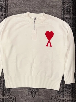Vit stickad tröja från Ami Paris - Vit stickad tröja från Ami Paris med rund halsringning och ribbade muddar. På bröstet finns ett rött hjärta och bokstaven 'A' som signaturdetalj. Tröjan har en klassisk passform och är perfekt för en stilren look.