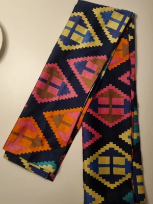 Färgglad halsduk  - Säljer en lång halsduk i mjukt tyg med färgstarkt geometriskt mönster i blått, gult, orange, rosa och turkost. Basen är mörkblå och mönstret består av trianglar och rutor i olika färger. Perfekt för dig som vill sticka ut och addera färg till din outfit.