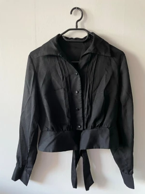 Svart blus med knytband  - Stilren svart blus med långa ärmar, bred krage och knytband i midjan. Framtill har den dekorativa veck och knappar. Gjord i ett genomskinligt mesh-liknade tyg.
