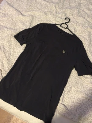 Svart t-shirt med liten logga - En enkel svart t-shirt med rund hals och korta ärmar. På bröstet sitter en liten logga i form av ett märke. T-shirten har en klassisk passform och är tillverkad i mjukt bomullstyg, perfekt för en clean och stilren look.