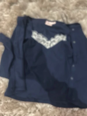 Mörkblå pyjamas med spetsdetalj - Snygg mörkblå kofta med knappar framtill och vit spetsdetalj över bröstet. Mjuk och bekväm modell med lång ärm, perfekt för mysiga kvällar hemma. Klassisk design med en feminin touch tack vare spetsen.