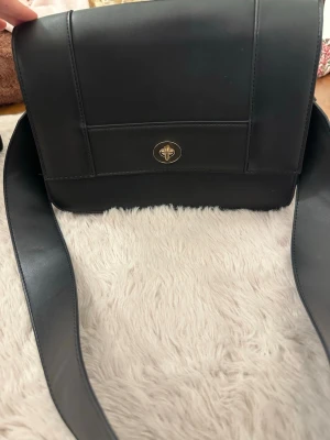 Svart axelväska med bred rem - Stilren svart axelväska i slätt konstläder med bred axelrem och guldfärgat vridlås framtill. Väskan har en rektangulär form och minimalistisk design som passar till många olika outfits. Perfekt för dig som gillar klassiska accesso soppar.