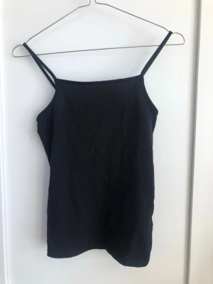 Svart enkelt linne - Snygg svart linne med smala axelband och rak halsringning. Perfekt att styla lager på lager eller bära som den är. Tillverkad i mjukt bomullsmaterial för en bekväm känsla.