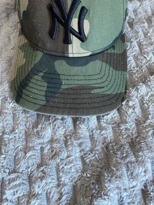 NY Yankees camo keps från New Era - Säljer en grön camo keps från New Era, modell 9FORTY, med broderad svart NY Yankees-logga framtill. Kepsen har böjd skärm och justerbar rem bak. Tillverkad i bomullsmix och har klassisk streetwear-vibe.