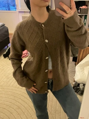 Brun stickad kofta med pärlknappar - Mysig brun stickad kofta med diskret mönster och fina pärlknappar. Koftan har rund halsringning, långa ärmar och en avslappnad passform. Perfekt att slänga över en t-shirt för en chill look.