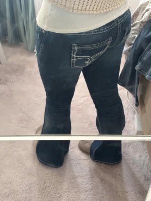 Mörkblå bootcut jeans med kontrastsömmar - Säljer ett par mörkblå bootcut jeans med vita kontrastsömmar och snygg detalj på bakfickan. Jeansen har slitningar framtill och en klassisk femficksmodell. Perfekta för dig som gillar en avslappnad men trendig stil. Hör gärna av er för fler bilder💘pris går att diskutera