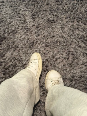 Vita Adidas Stan Smith sneakers - Klassiska vita Adidas Stan Smith sneakers i läder med perforerade ränder på sidorna och grå detaljer på hälen. Skorna har rund tå, vita snören och låg profil. Perfekta för en clean och tidlös stil.