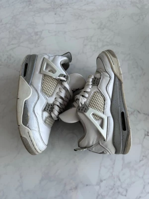 Vita Air Jordan 4 Retro sneakers - Säljer ett par vita Air Jordan 4 Retro sneakers med grå och svarta detaljer samt röd Jumpman-logga på plösen. Skorna har meshpaneler, klassiska plastdetaljer och tjock sula. Tillverkade i läder och mesh för en ikonisk look.