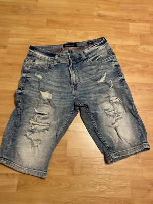 Blå slitna jeansshorts  - Säljer ett par blå jeansshorts  med slitna detaljer och längre ben. Klassisk femficksmodell med dragkedja och knapp. Perfekta för en avslappnad stil i sommar. Materialet är bomull och shortsen har en normal passform. 