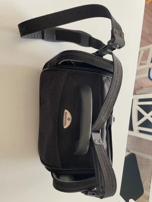 Svart axelväska från Samsonite - Snygg svart axelväska från Samsonite med justerbar axelrem och robust handtag. Väskan har flera fack med dragkedja och är tillverkad i slitstarkt tygmaterial med detaljer i plast. Perfekt för dig som vill ha en praktisk och stilren väska till vardagen.