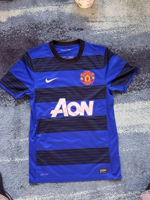  Manchester United tröja Nike 2011-2013 äkta.  - Snygg blå Manchester United matchtröja från Nike med svarta horisontella ränder och klubbmärke på bröstet. Tröjan har vit Nike-logga och AON som sponsortryck. Tillverkad i Dri-FIT material som andas, perfekt för träning eller match.
