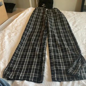 Svarta vida rutiga byxor - Säljer ett par svarta byxor med vitt rutmönster och vida ben. Byxorna har hög midja och stängs med knapp framtill. Perfekta för dig som gillar en loose och relaxed stil.Perfekt till vår/sommar.
