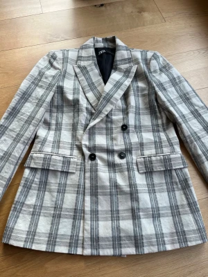 Rutig kavaj från Zara i grått och vitt - Snygg dubbelknäppt kavaj från Zara med grå och vit rutig design. Klassisk krage, svarta knappar och två fickor med lock framtill. Perfekt för dig som vill ha en trendig och stilren look. Tunn fodring och rak passform.