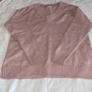 Ljusrosa stickad tröja med v-ringning - Mysig ljusrosa stickad tröja med v-ringad hals och långa ärmar. Tröjan har bred ribbstickad mudd vid ärmslut och nederkant. Perfekt för lager på lager och ger en avslappnad men ändå stilren look.