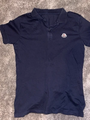 Mörkblå Moncler t-shirt med krage - Snygg mörkblå piké från Moncler med klassisk krage och knappslå framtill. Liten Moncler-logga på bröstet. Perfekt till jeans eller shorts för en clean och stilren look.