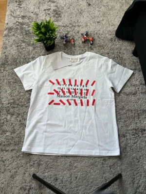 Vit Maison Margiela t-shirt med print - Vit t-shirt från Maison Margiela med kort ärm och rund hals. Framsidan har ett unikt svart och rött tryck med märkets logotyp och siffror. Tillverkad i mjuk bomull, perfekt för en trendig och avslappnad look.