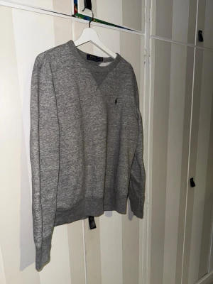 Grå sweatshirt från Polo Ralph Lauren - Klassisk grå sweatshirt från Polo Ralph Lauren med rund halsringning och diskret broderad logga på bröstet. Tröjan har ribbade muddar vid ärmslut och nederkant, perfekt för en avslappnad och stilren look.