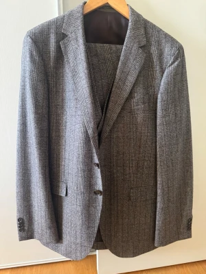  Boss Grå rutig 3-delad  kostym - Snygg grå rutig 3-delad kostym i 100% ull. Kavajen har två knappar, smala slag och fickor med lock. Kostymen har ett stilrent mönster och passar perfekt till dig som vill ha en tidlös look.