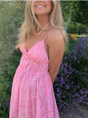Rosa långklänning med zebramönster - Söt rosa långklänning med vitt zebramönster och smala axelband. Klänningen har v-ringning och en luftig, flowy passform som ger en avslappnad och somrig känsla. Perfekt för varma dagar och trädgårdshäng.